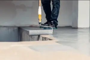 A person applies gray epoxy resin with a roller on a concrete floor, possibly preparing for polyaspartic garage flooring. Wearing dark pants and protective footwear, they focus intently on the task amid the bustling construction or renovation scene.
